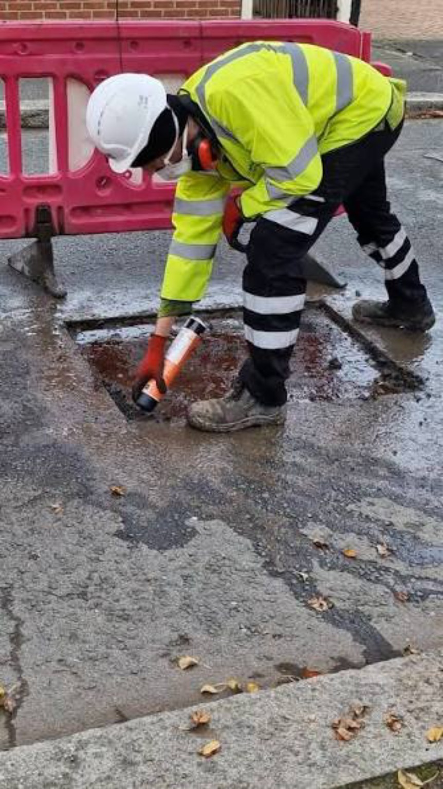 Worker attending to a local patch in the road surface during reinstatement work in Fife, with barriers behind