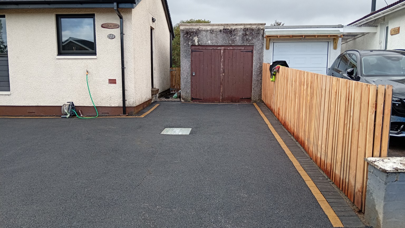 Finished Fort William tarmac driveway leading to the garage with charcoal and buff block edging beside the timber fence