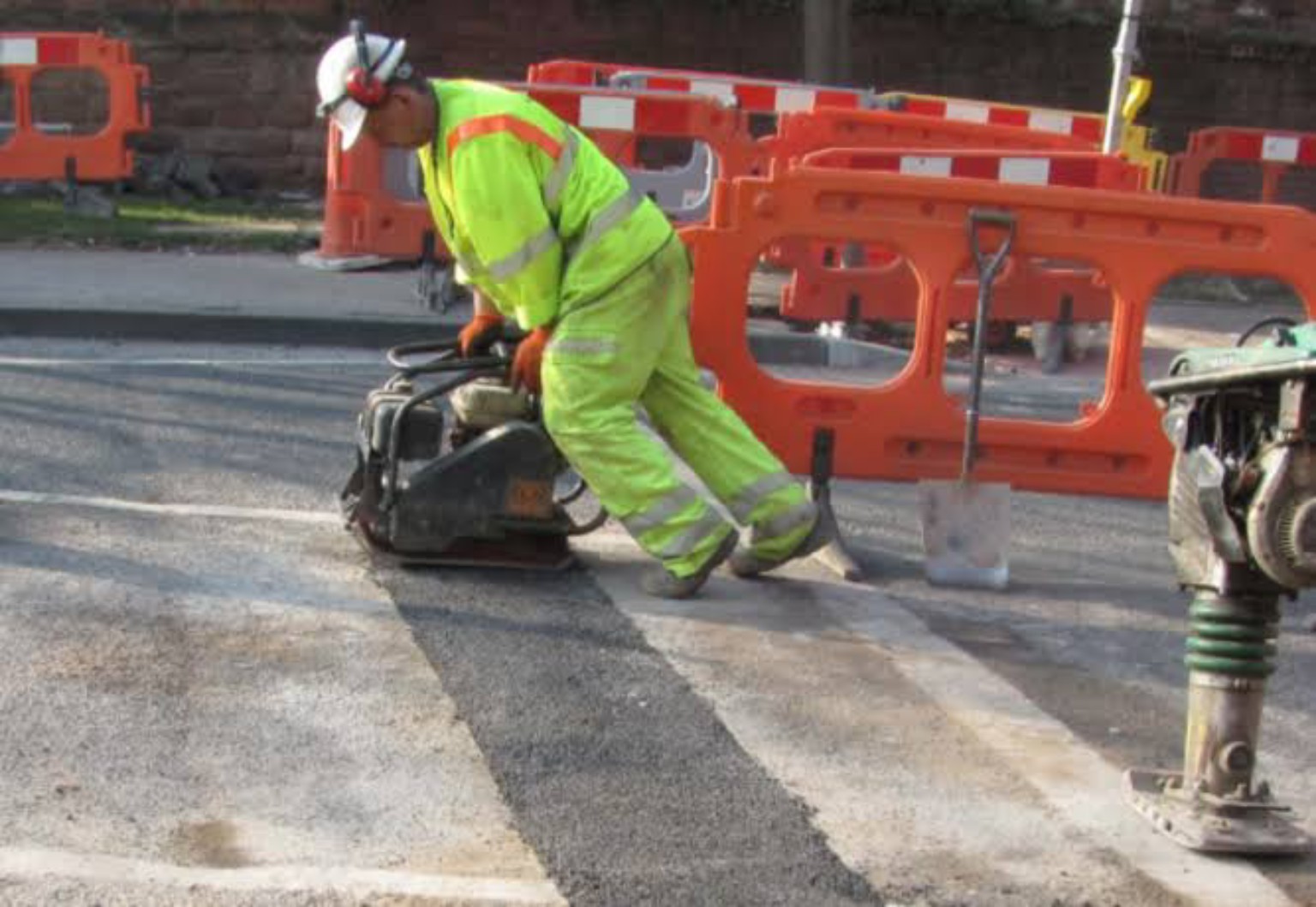 Worker using a plate compactor on a narrow tarmac reinstatement in Glasgow, beside safety barriers