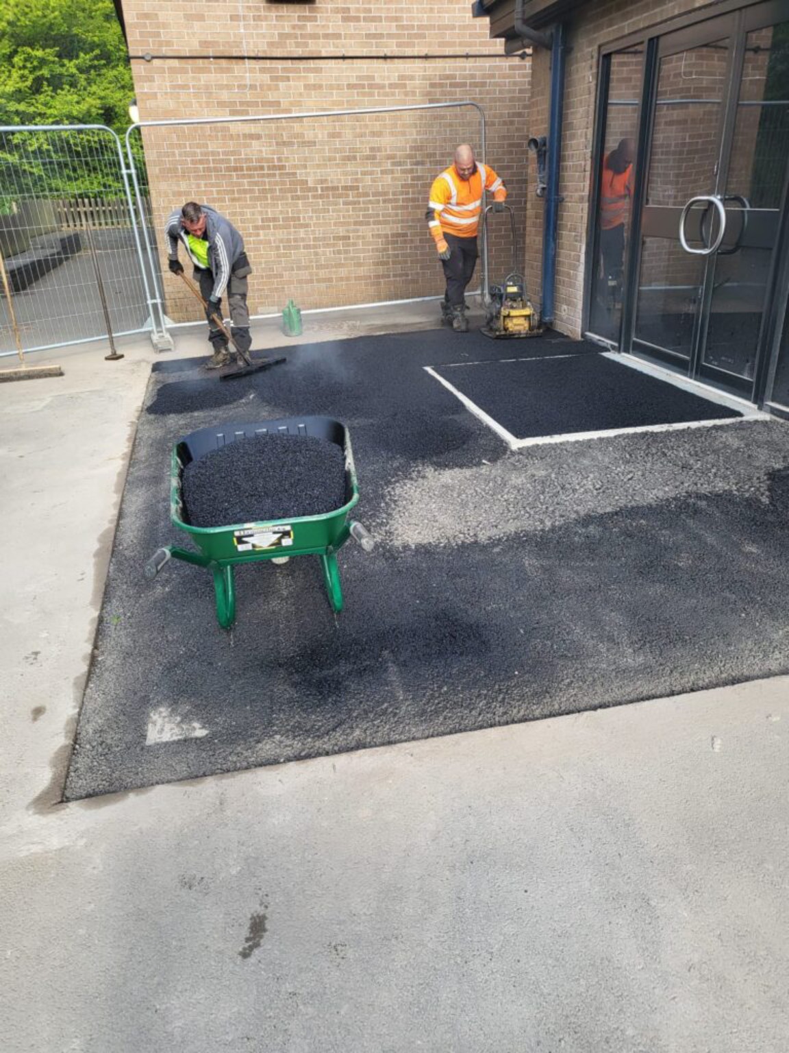 Workers hand laying and compacting hot tarmac at a school entrance in Gourock, Inverclyde, with a wheelbarrow and plate compactor