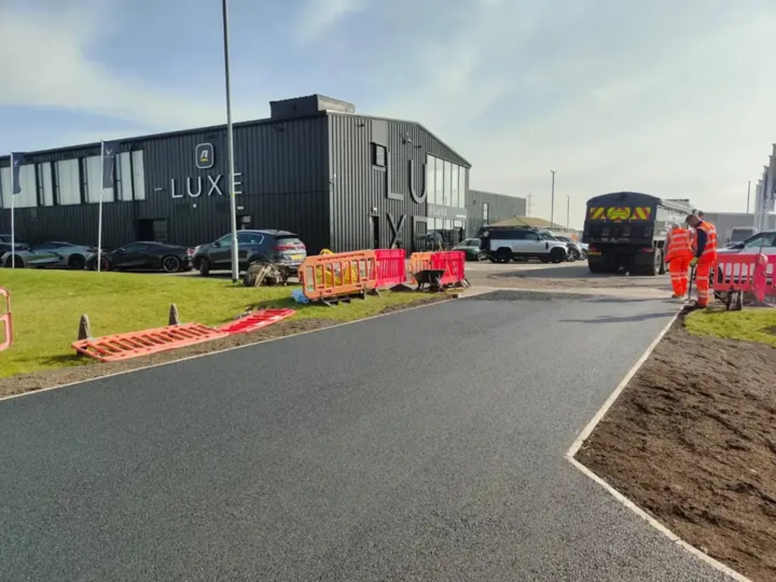 Fresh tarmac access road at a retail park with kerbed edges, barriers and workers finishing the surface