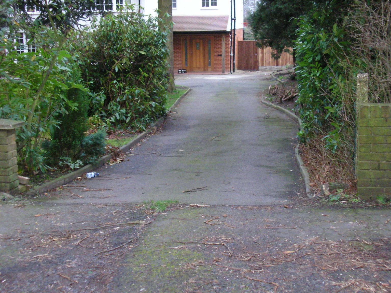 Worn tarmac driveway between brick entrance pillars and mature shrubs before resurfacing