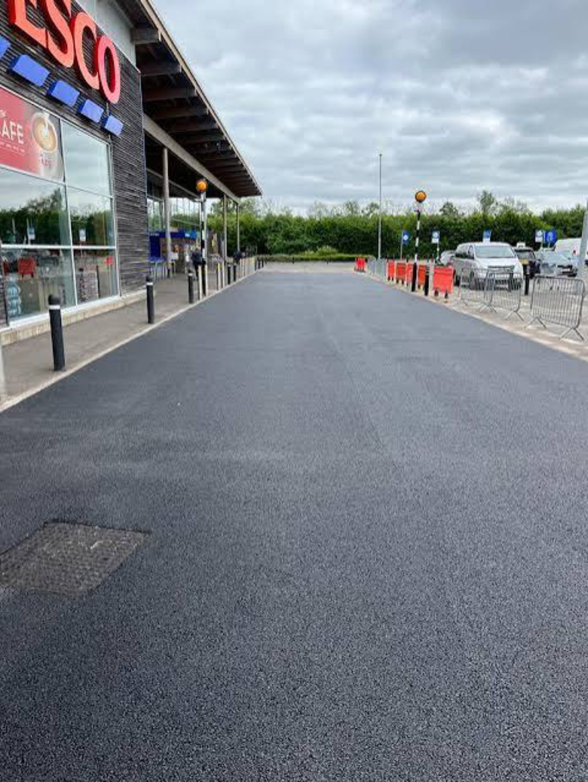 Freshly resurfaced tarmac beside a Tesco store entrance with pedestrian crossing posts and barriers at the edge of the work area