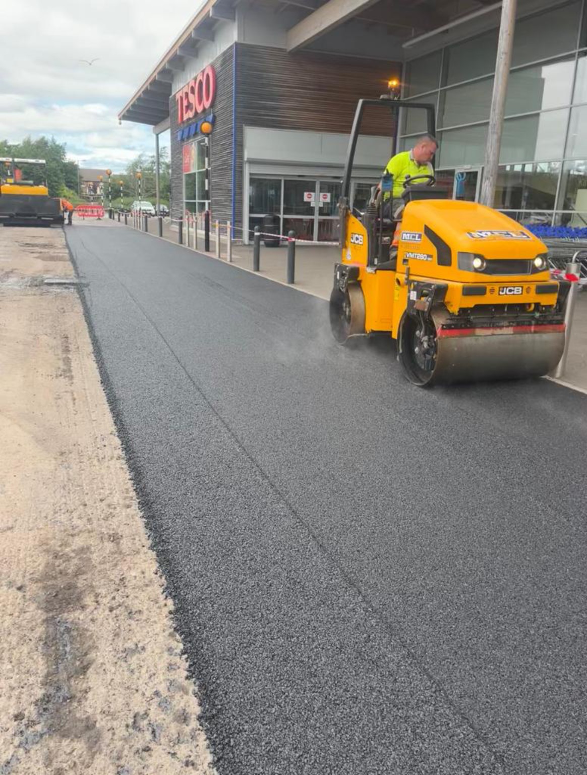Roller compacting new tarmac outside a Tesco store entrance during resurfacing